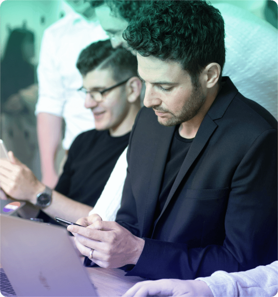 A group of people sit closely together, focused on their smartphones and a laptop. The atmosphere is collaborative and engaged, with a hint of concentration.
